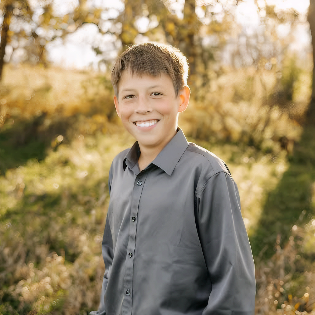 portrait of young boy standing in a field surrounded by trees smiling at camera